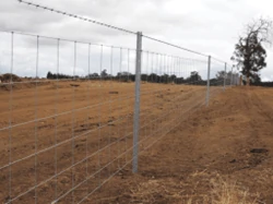 A construction worker builds a fence in a grassy field under a clear blue sky.